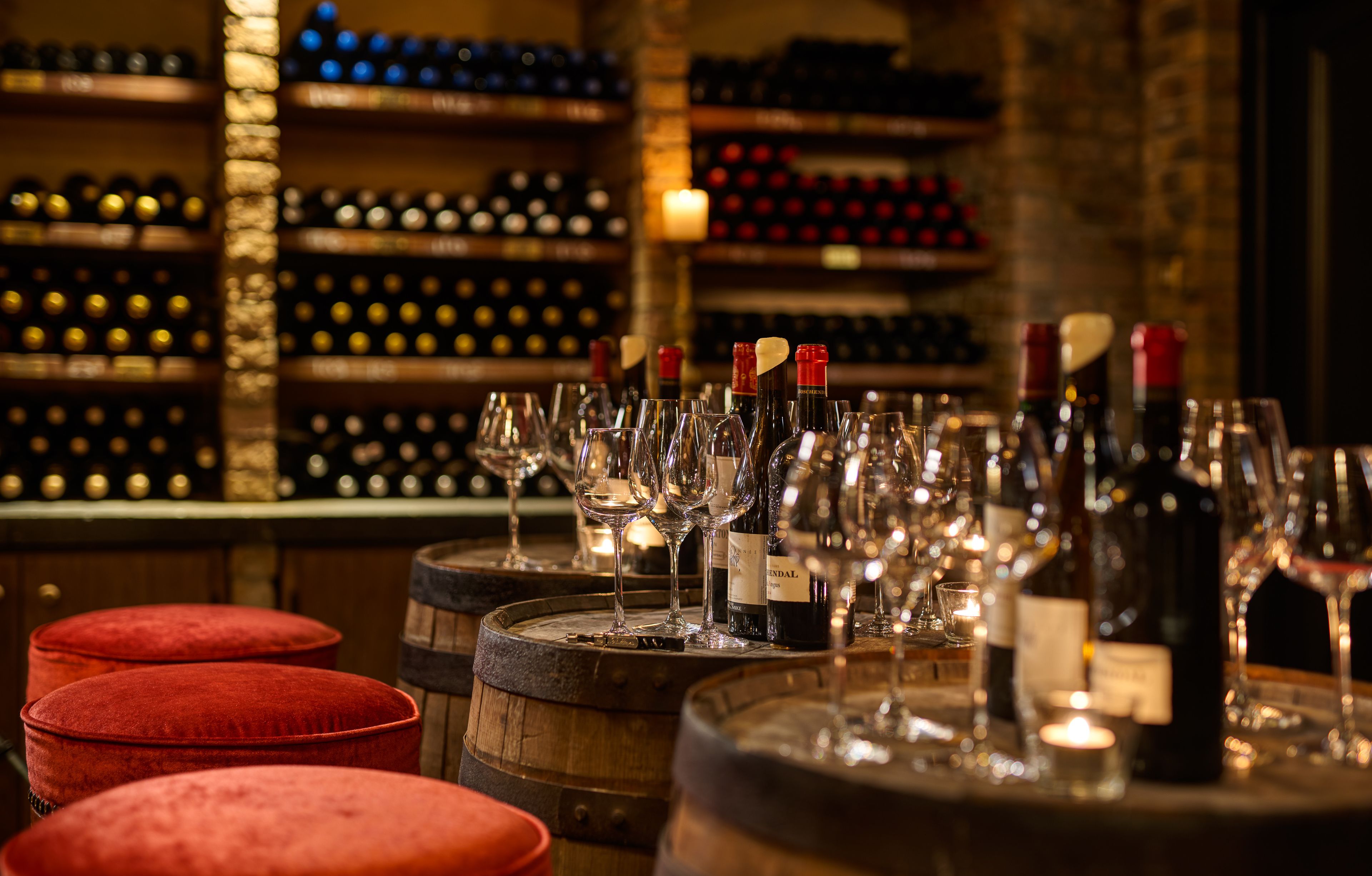 A bar area with wine bottles and glasses on wooden barrels, red cushioned stools, and shelves with wine bottles in the background.