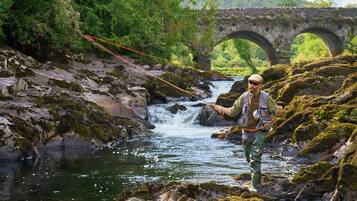 Un hombre pesca junto a un río, con un puente de piedra al fondo.