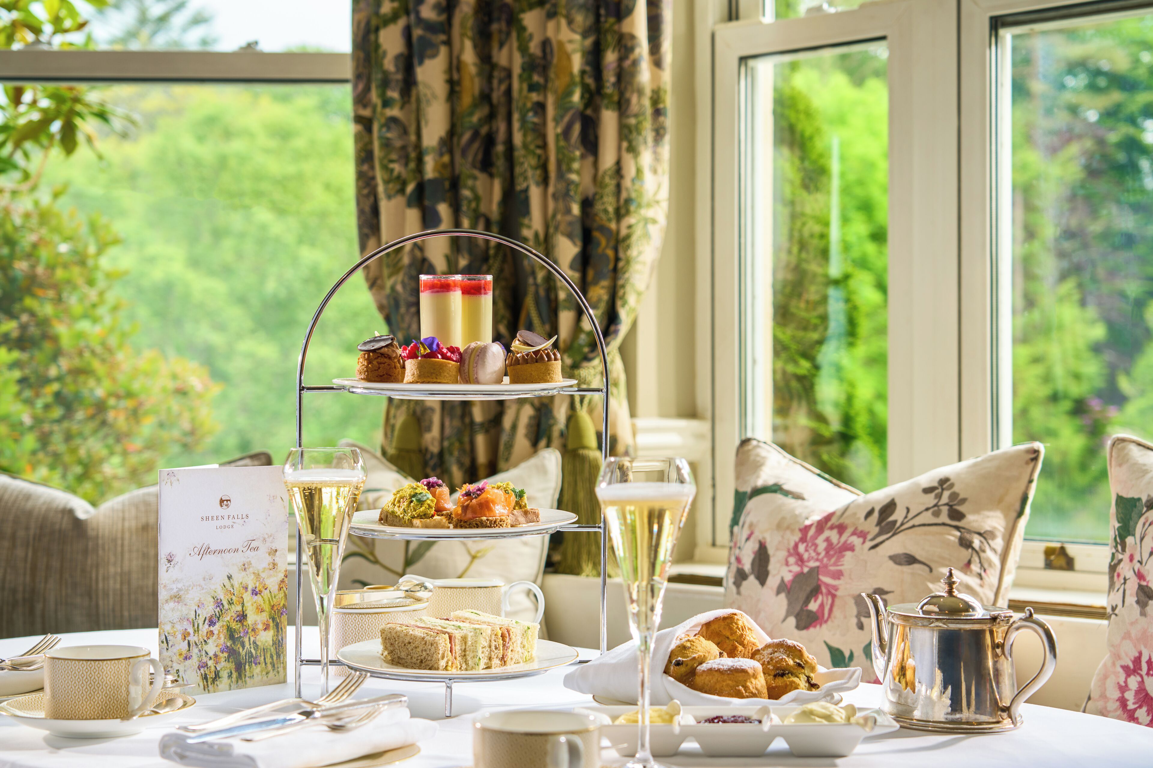 A three-tiered dessert stand with various pastries and a glass of champagne, set on a table with a tea menu, cups, and a silver teapot.