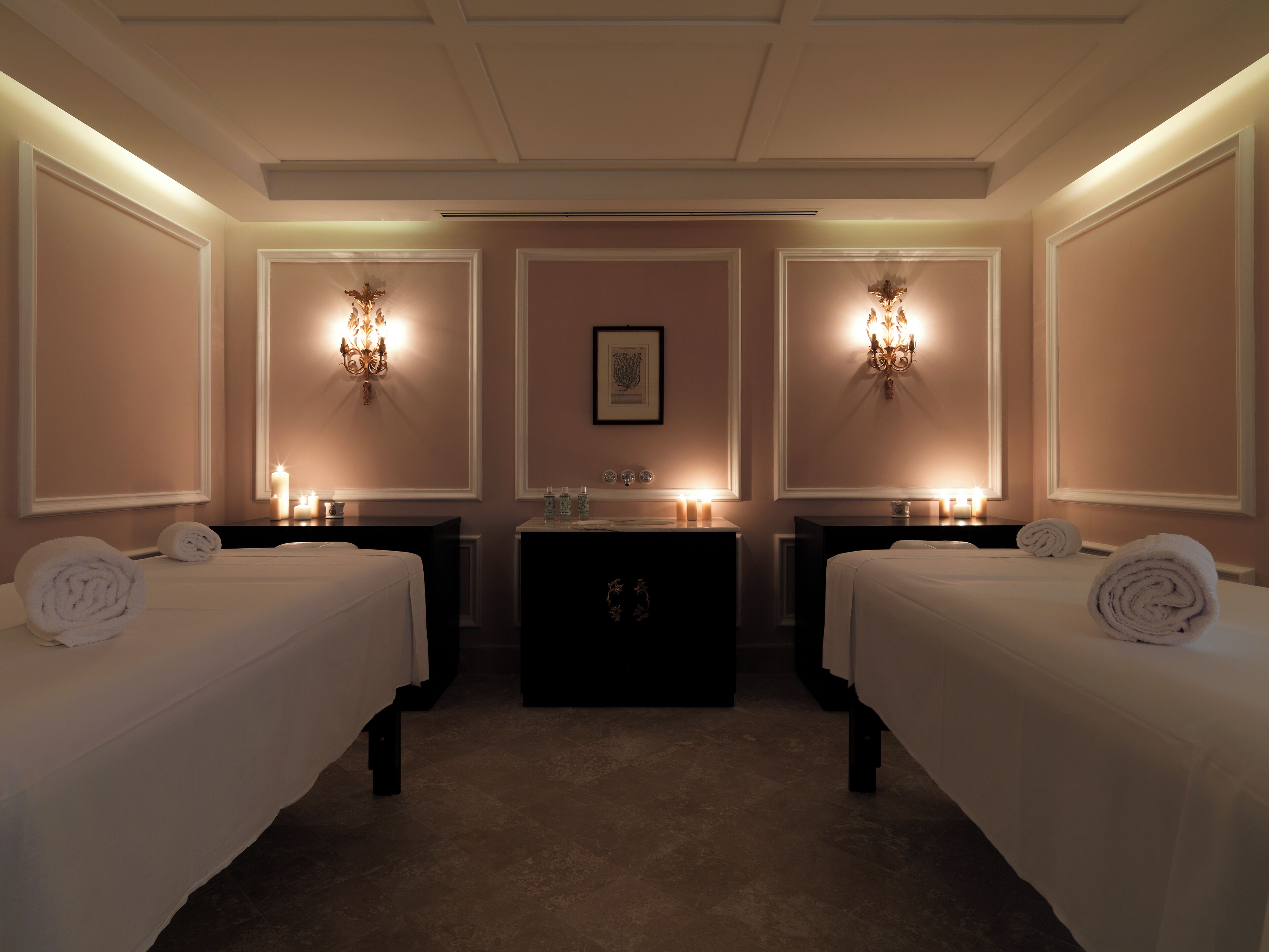 A hotel room with two beds, a central table, and wall-mounted sconces.