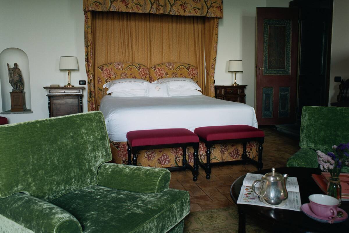 A hotel room with a large bed, green upholstered chairs, a small table with a teapot and cup, and a door with intricate woodwork.