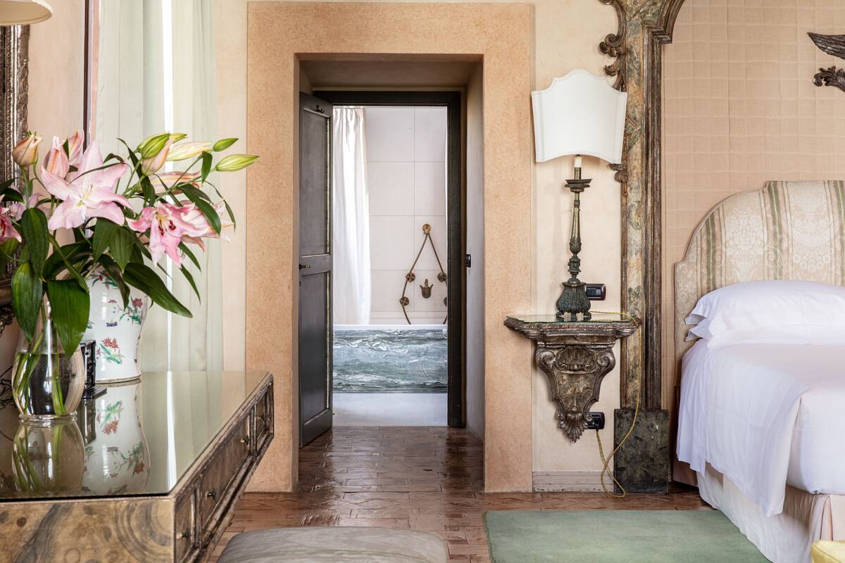 A hotel room with a bed, a nightstand, a mirror, a vase with flowers, and a view of a beach through an open door.