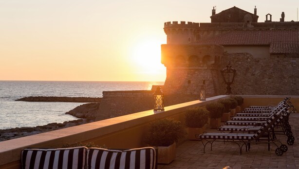 A sunset view from a balcony with a stone wall, a row of sun loungers, and a building with a tower. - La Posta Vecchia Hotel (Ladispoli)