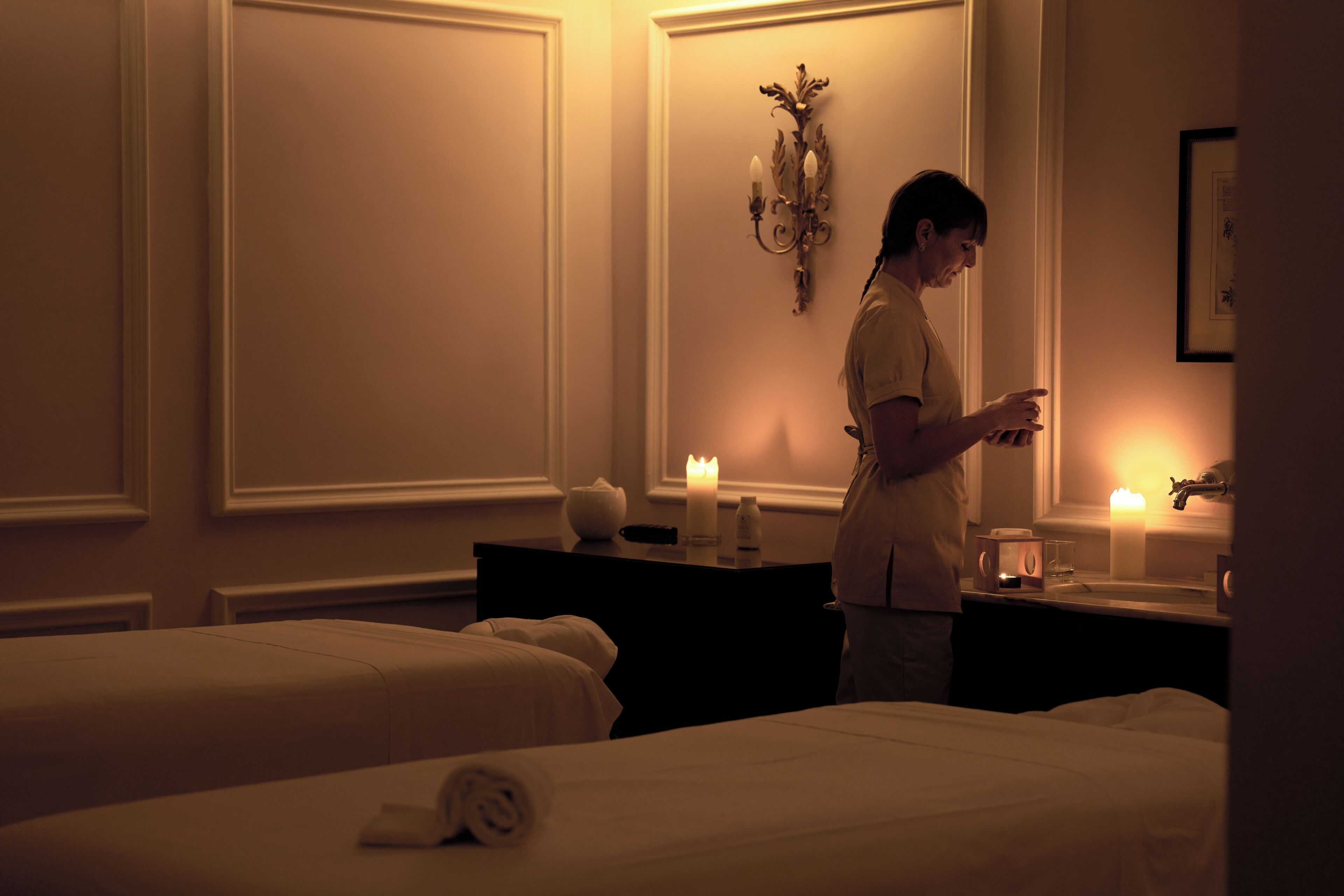 A hotel room with two beds, a nightstand with a candle, a wall-mounted lamp, and a framed picture.