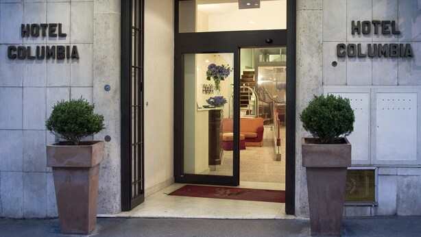 The entrance of Hotel Columbia with a glass door, a red carpet, and two potted plants. - Hotel Columbia (Rome)