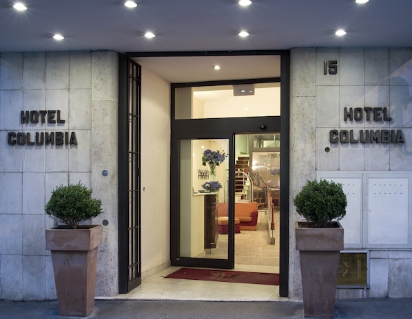 The entrance of Hotel Columbia with a glass door, a red carpet, and two potted plants. - Hotel Columbia (Rome)