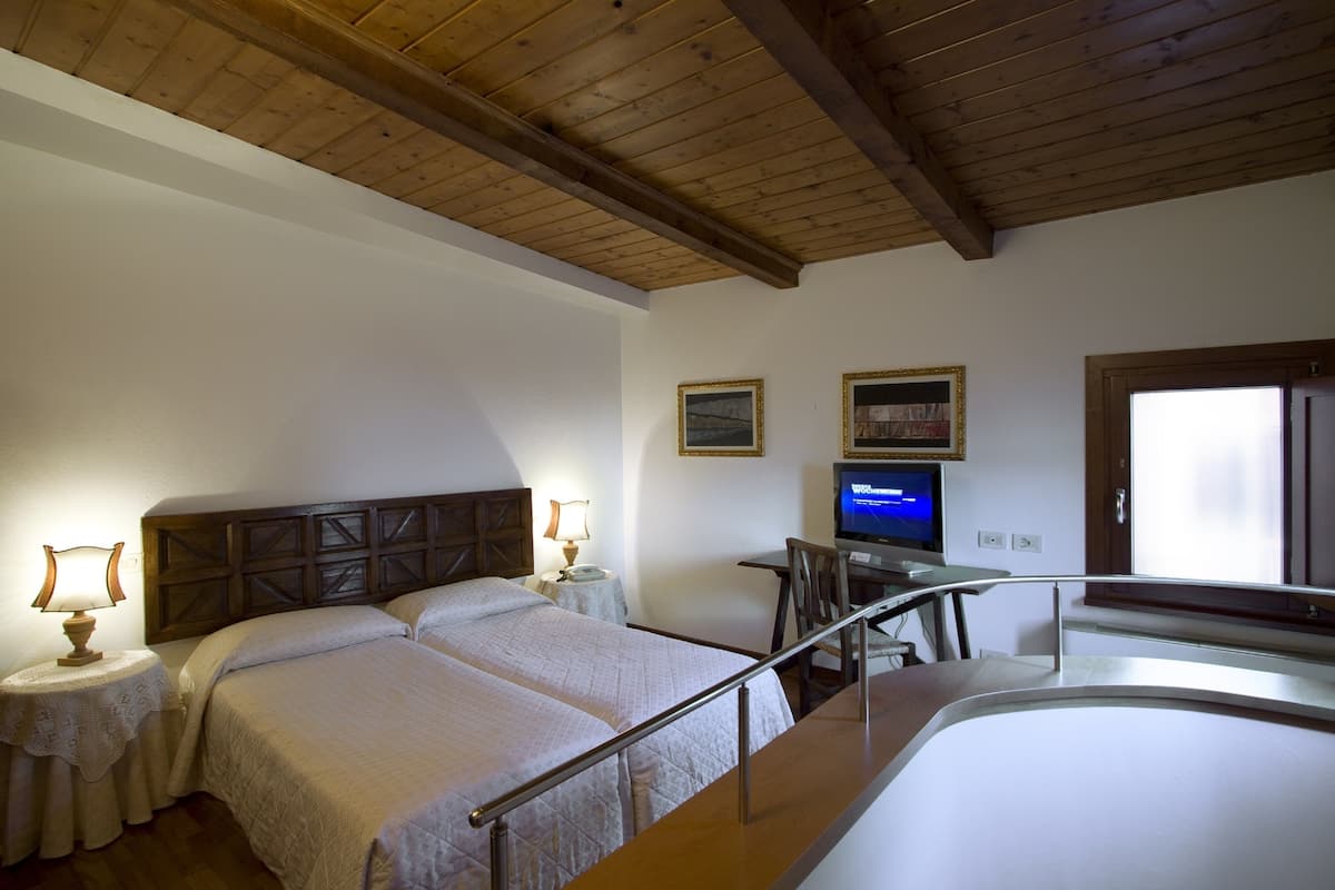 A hotel room with a wooden ceiling, a bed with a headboard, a desk with a computer, a TV, and a window.