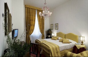 A hotel room with a bed, bedside lamps, a chandelier, a television, and a window with curtains.