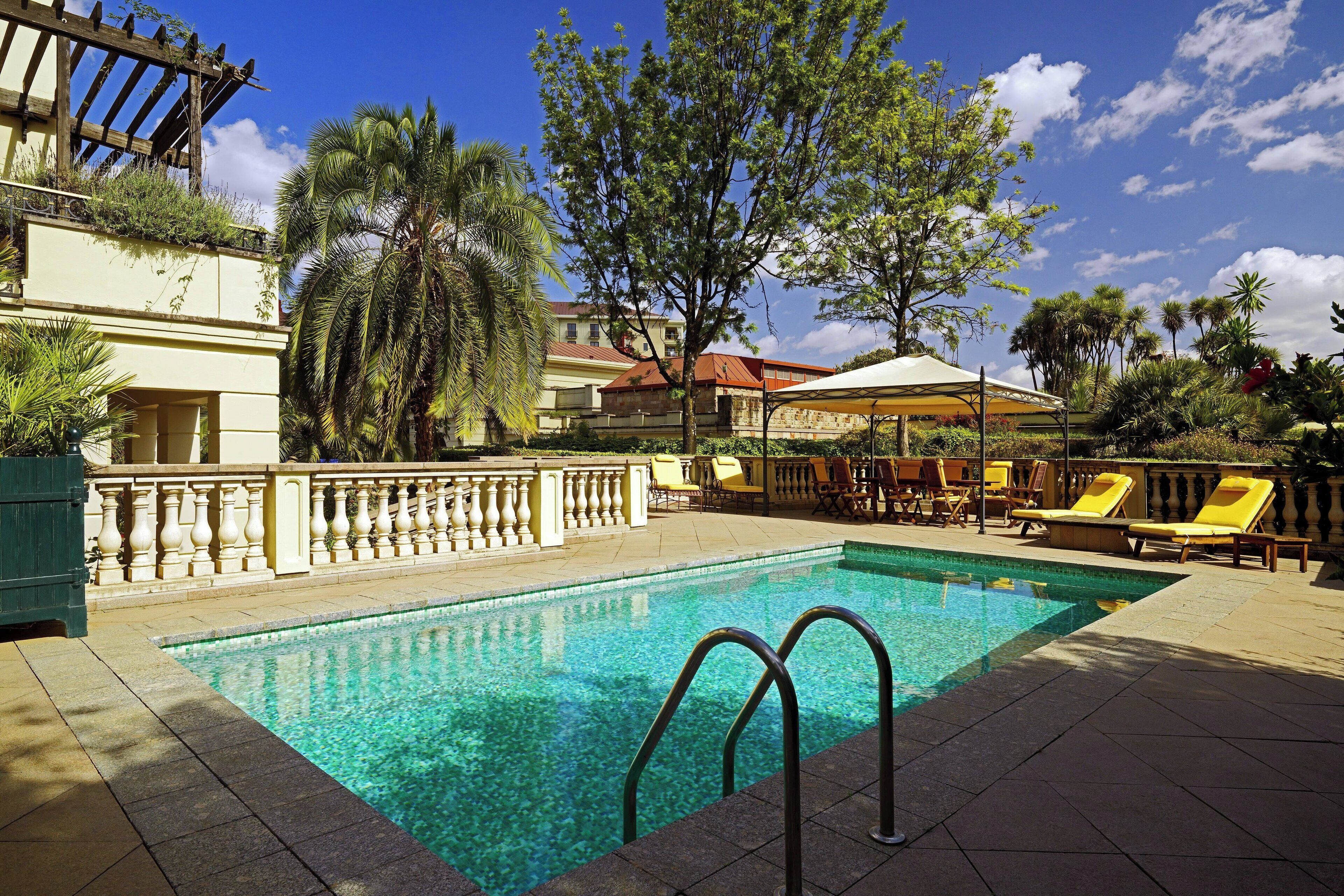 Ein Swimmingpool mit Liegestühlen und Sonnenschirmen, umgeben von Palmen und einem Gebäude mit einem Balkon.