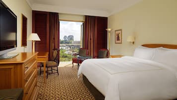 A hotel room with a large bed, a desk with a TV, a chair, a small table, and a balcony with a view of the city.