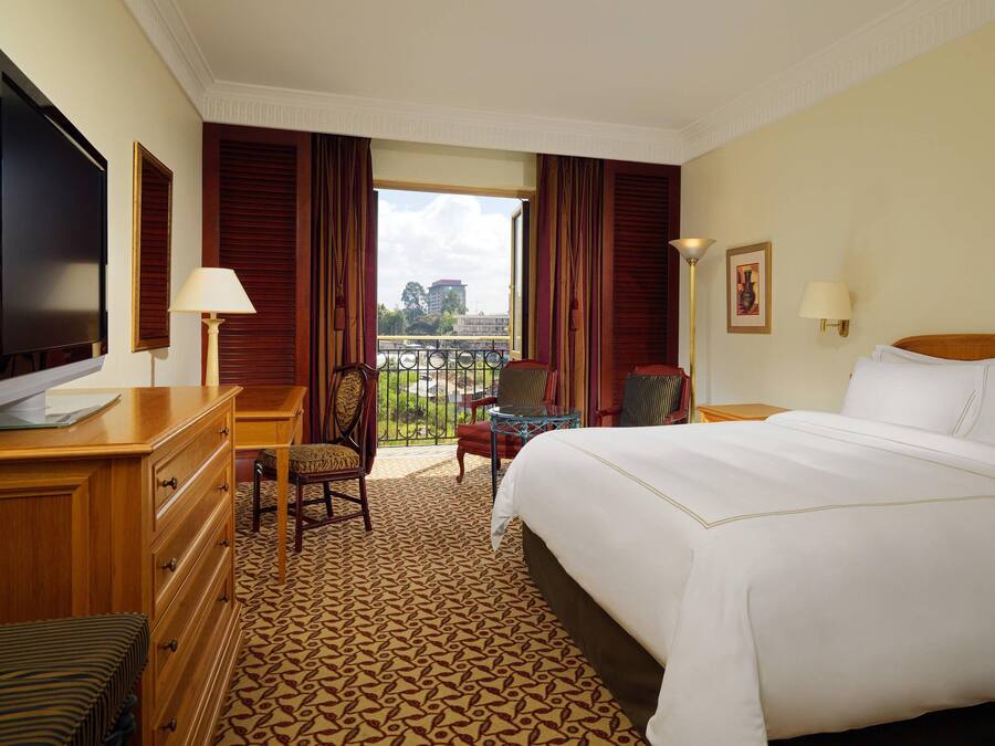 A hotel room with a large bed, a desk with a TV, a chair, a small table, and a balcony with a view of the city.