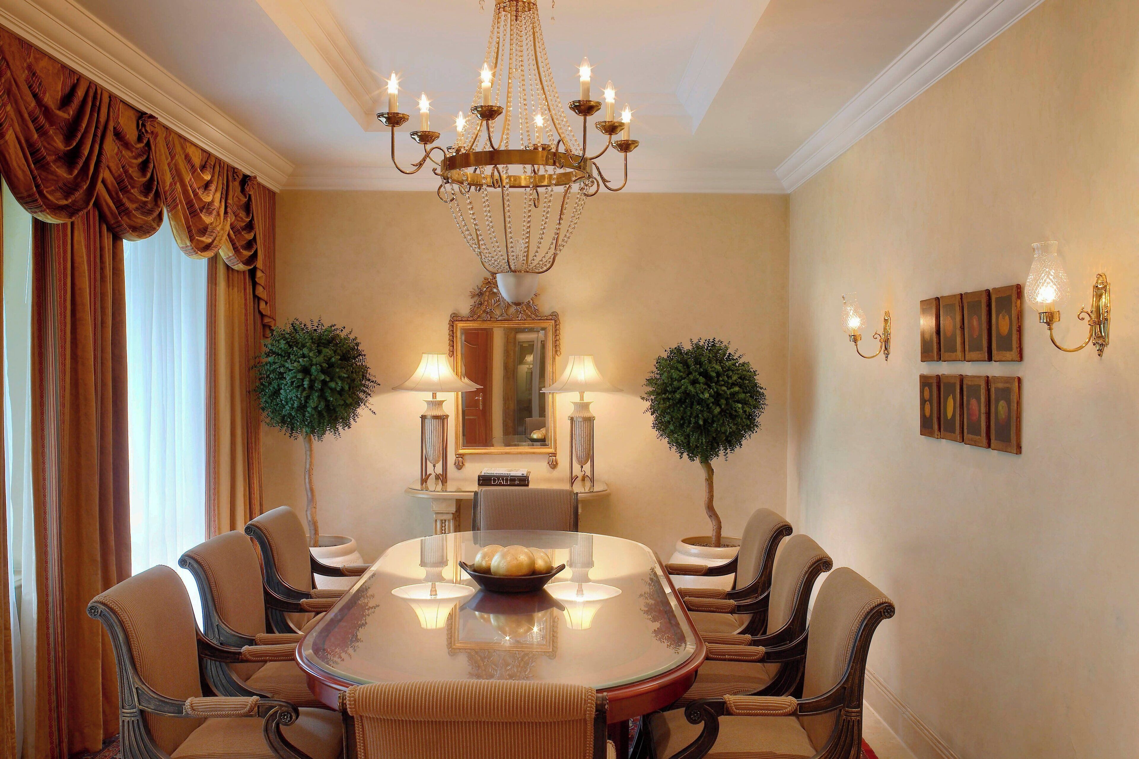 A formal dining room with a large oval table, six chairs, a chandelier, wall-mounted lamps, and framed pictures.
