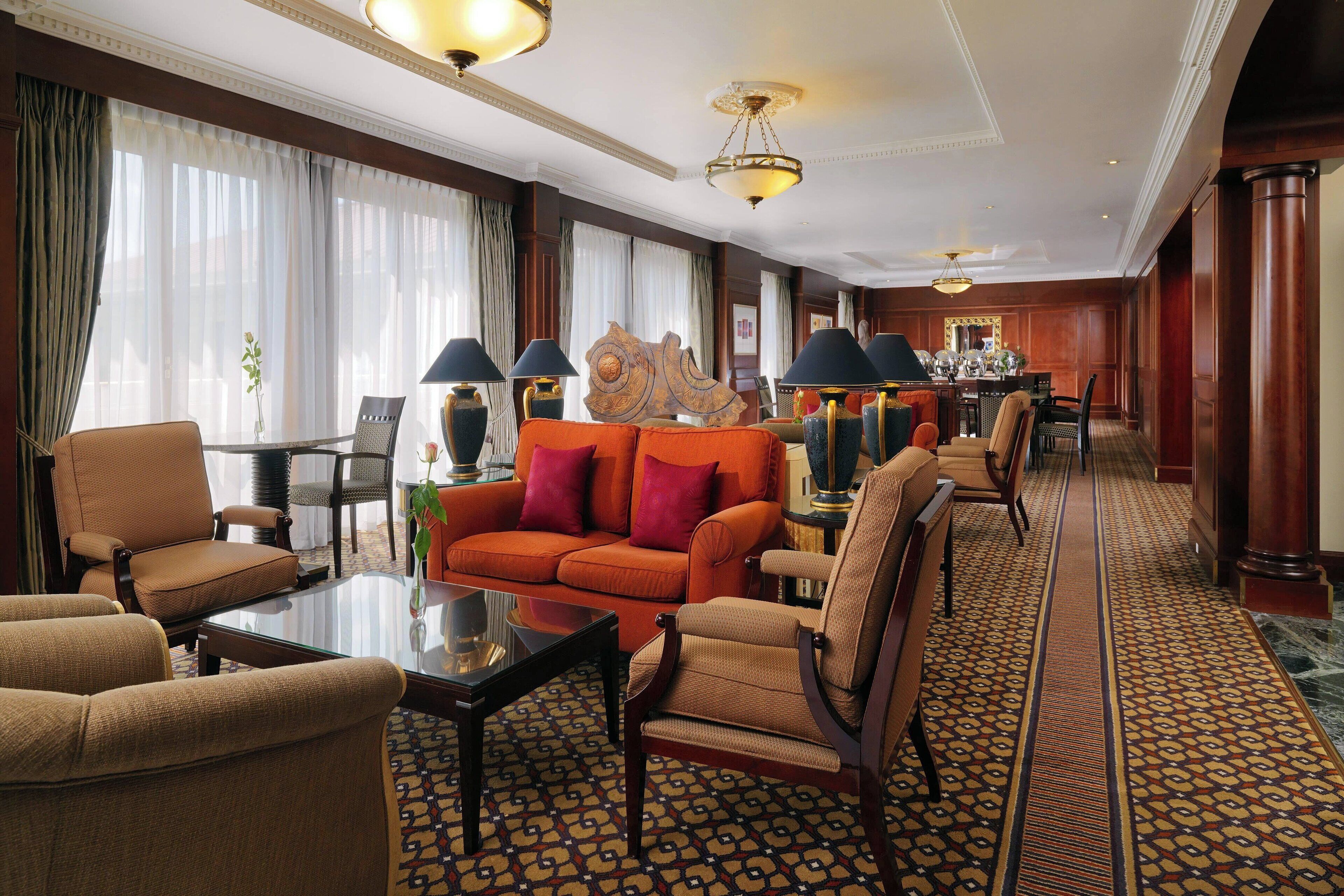 Eine geräumige Hotelhalle mit einem gemusterten Teppich, einer zentralen Sitzgruppe mit einer Couch und Sesseln sowie einem Essbereich im Hintergrund.