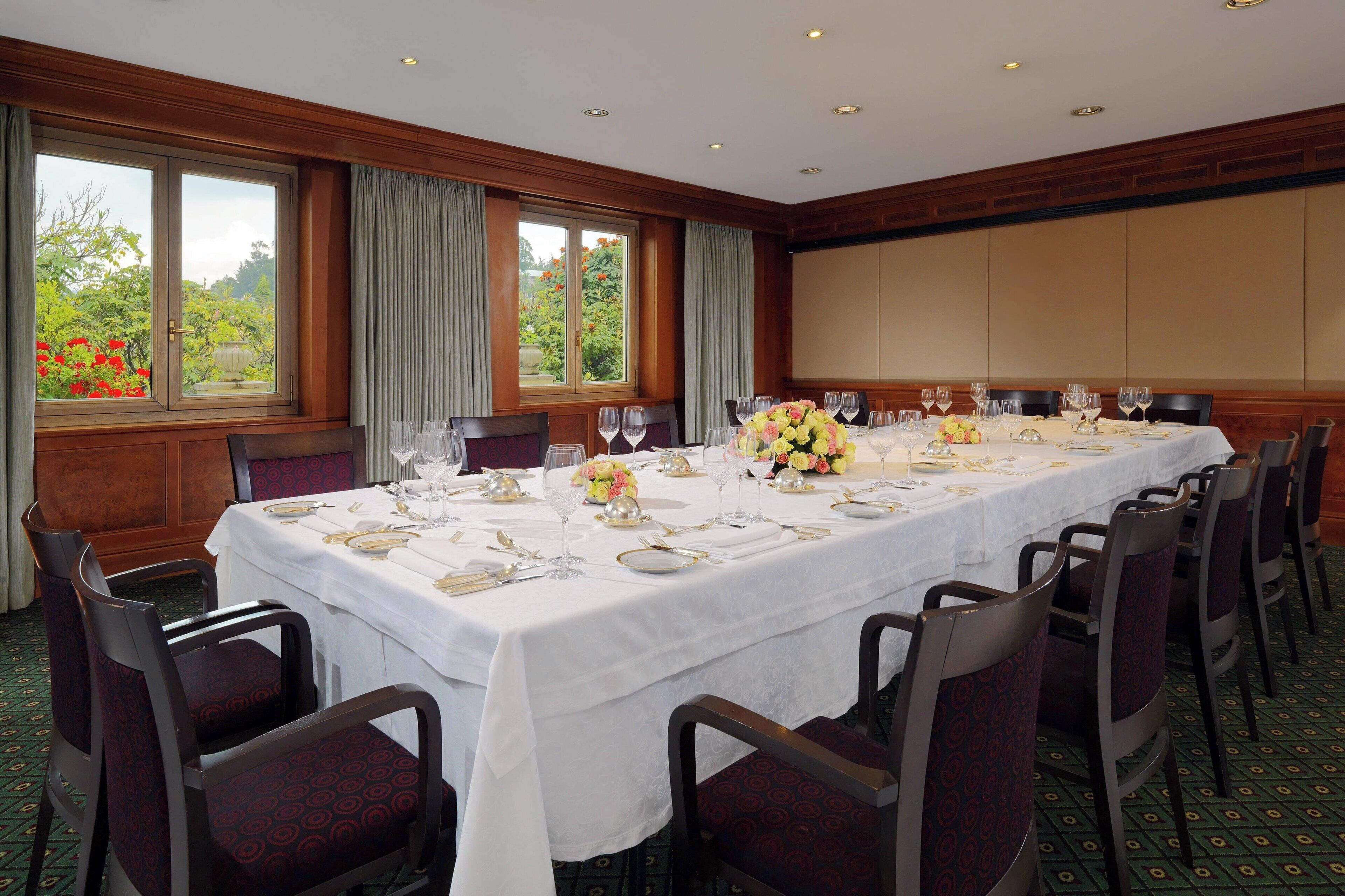A conference room with a long table set for a meeting, large windows with curtains, and a view of greenery outside.