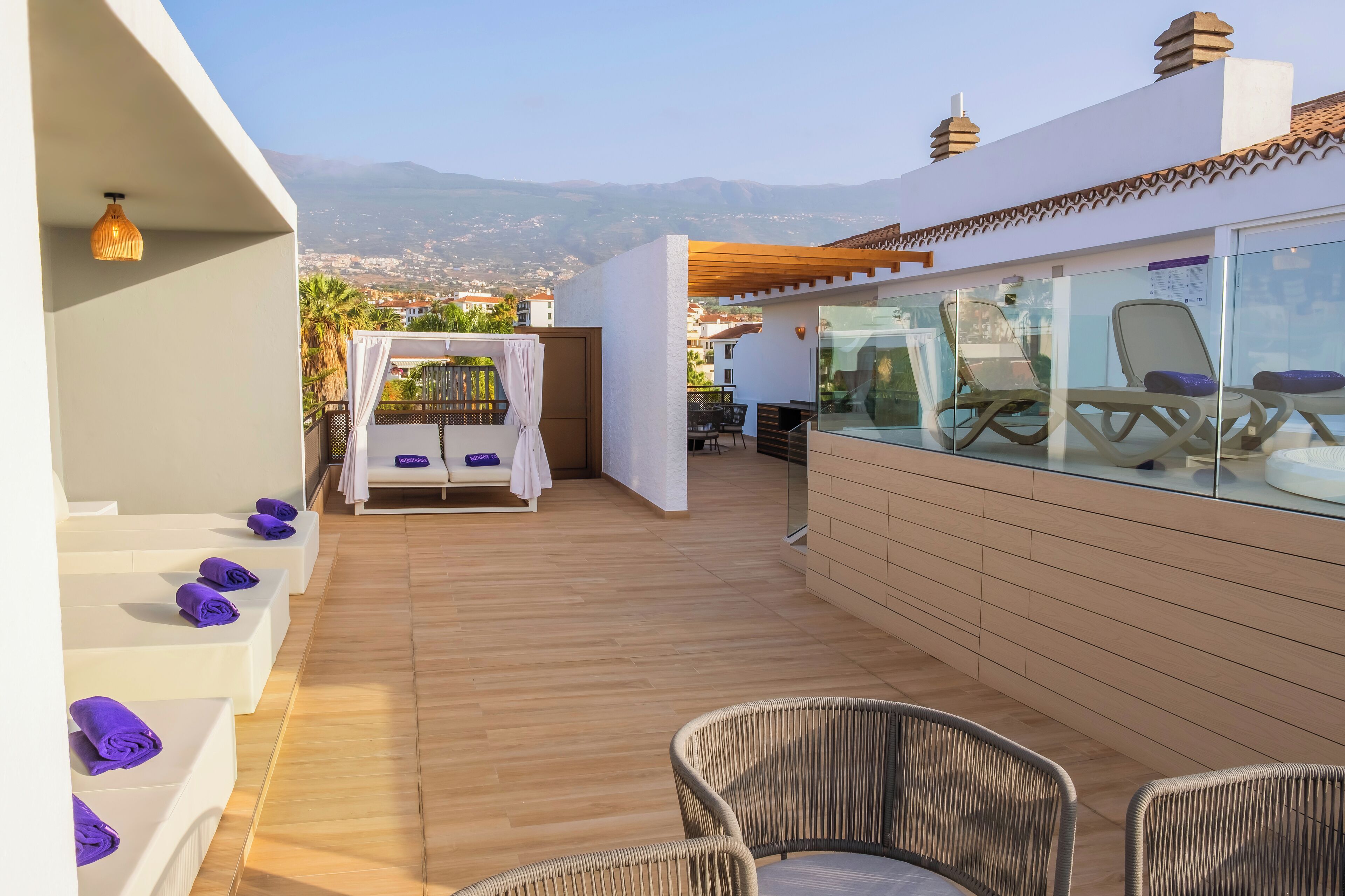 A rooftop terrace with wooden flooring, lounge chairs, and a glass wall offering a view of the city and mountains.
