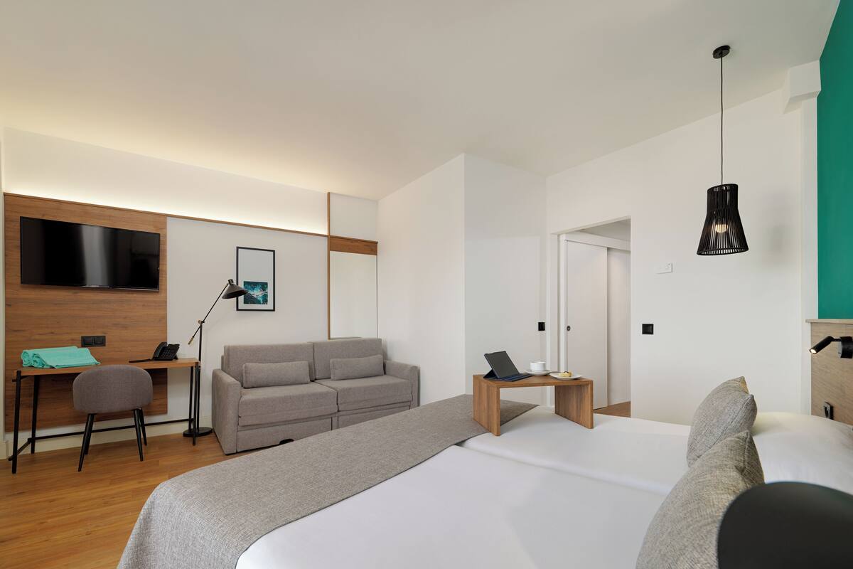 A modern hotel room with a large bed, a sofa, a desk with a computer, a TV, and a small table with a lamp.
