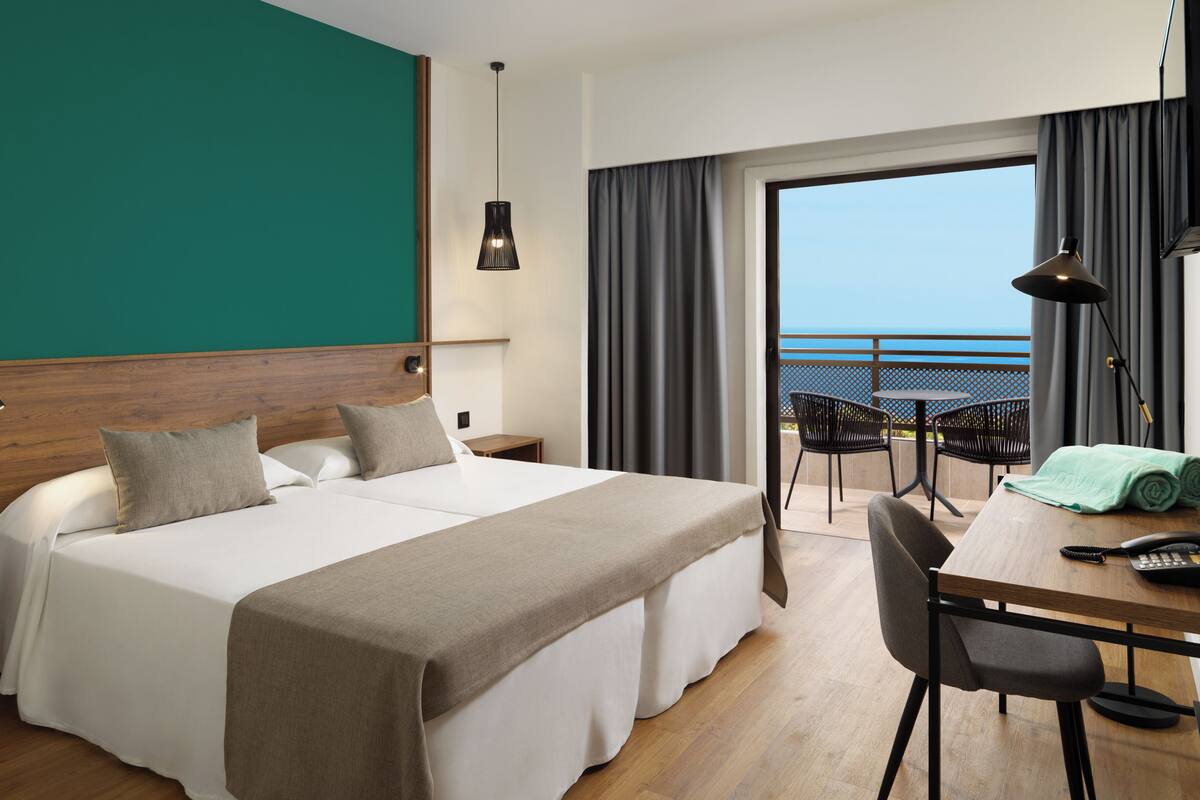 A hotel room with a large bed, a desk with a chair, a balcony with a view of the sea, and a window with curtains.
