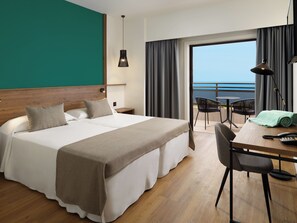 A hotel room with a large bed, a desk with a chair, a balcony with a view of the sea, and a window with curtains.