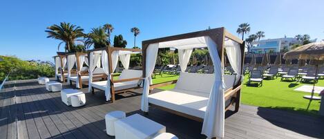 A rooftop deck with four wooden canopy beds, white cushions, and white cylindrical stools.