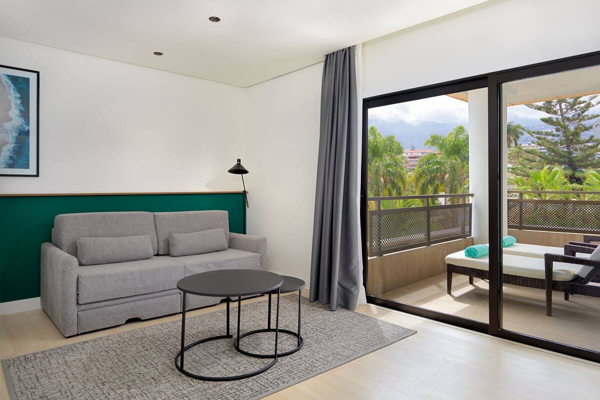 A modern living room with a grey sofa, a small round table, and a view of a balcony with outdoor furniture.