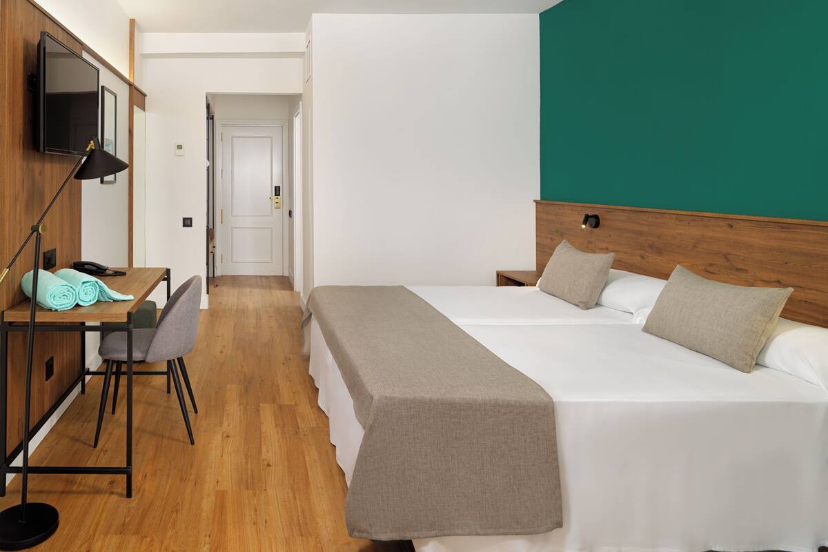 A hotel room with a wooden desk, a bed with two pillows, a bench, and a TV mounted on the wall.