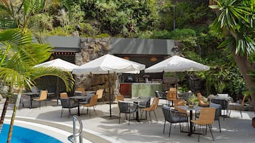Zona de piscina con mobiliario exterior, sombrillas y bar.