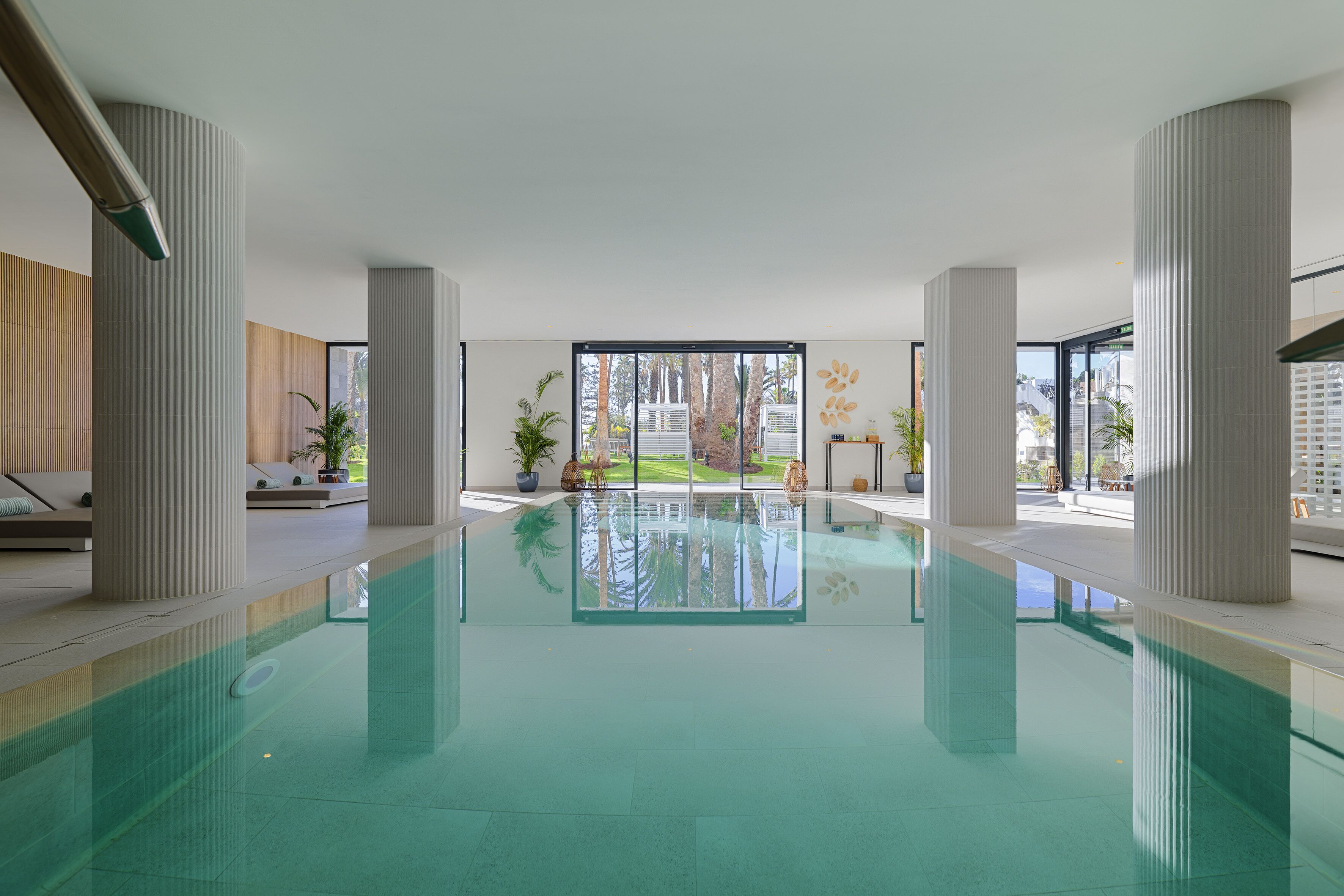 A modern indoor pool with clear glass walls, large pillars, and a view of a landscaped area outside.
