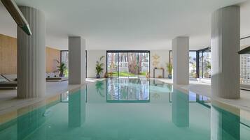 A modern indoor pool with clear glass walls, large pillars, and a view of a landscaped area outside.