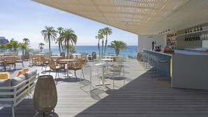 Zona de bar frente a la playa con mesas al aire libre, barra y vista al océano.