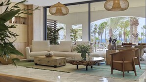 Una sala de estar moderna con un sofá, una mesa de centro y sillas, ventanas amplias con vistas a palmeras y al océano, y dos lámparas colgantes de mimbre.