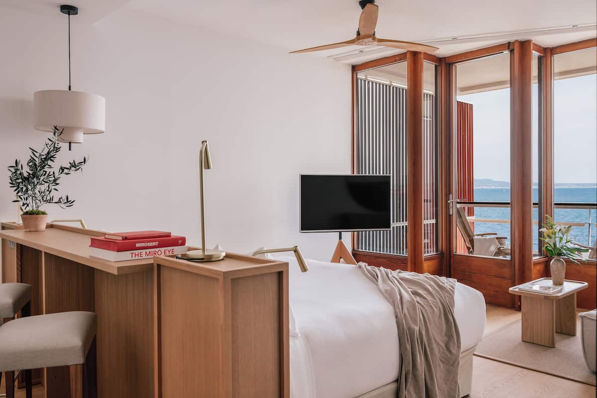 A modern bedroom with a bed, a desk with books, a TV, and a balcony with a view of the sea.