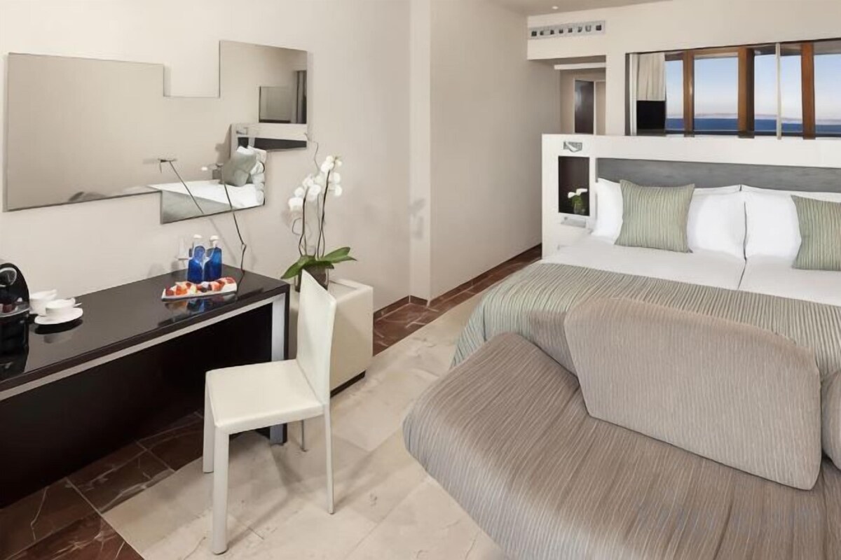 A hotel room with a bed, a desk, a chair, a mirror, and a view of the sea.