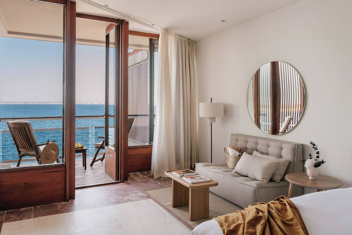 A hotel room with a balcony, a sofa, a bedside table, a lamp, and a large mirror.