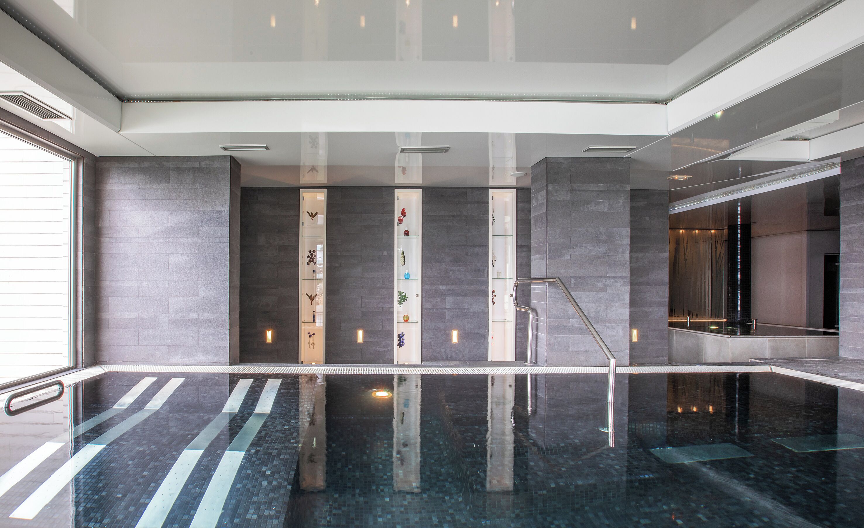 Une piscine intérieure moderne dotée d’un plancher en verre transparent, de murs en pierre grise et d’un escalier central.