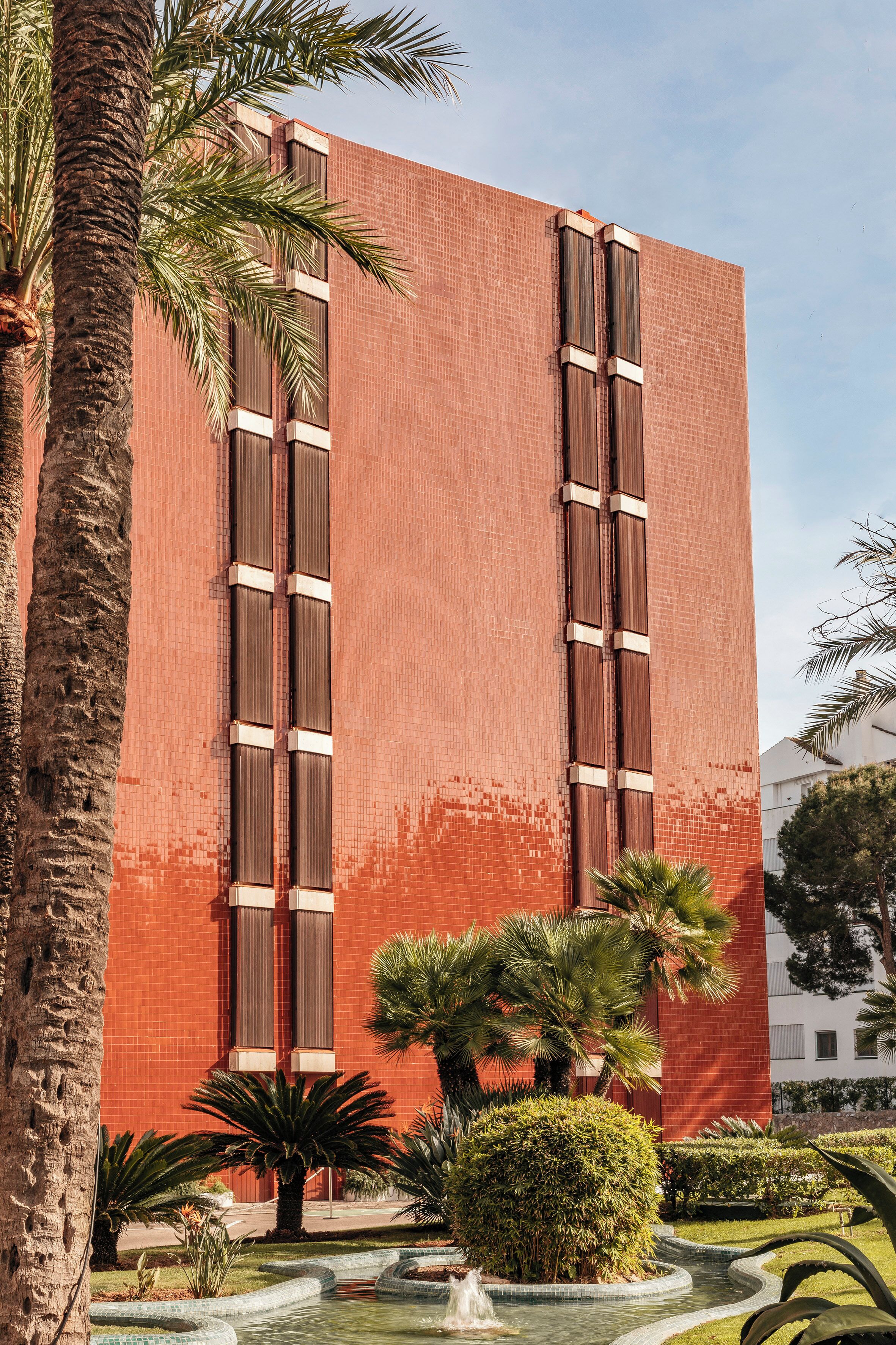 Un edificio de ladrillo rojo con ventanas verticales y un patio ajardinado con palmeras y una fuente.