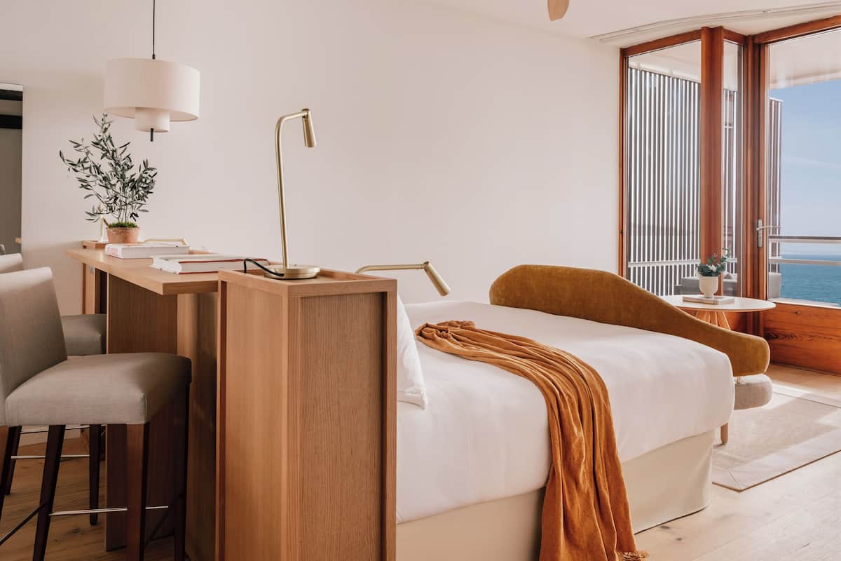 A modern hotel room with a wooden desk, a comfortable bed, a chair, and a view of the sea.