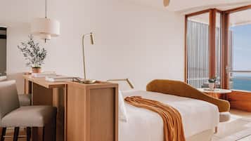 A modern hotel room with a wooden desk, a comfortable bed, a chair, and a view of the sea.