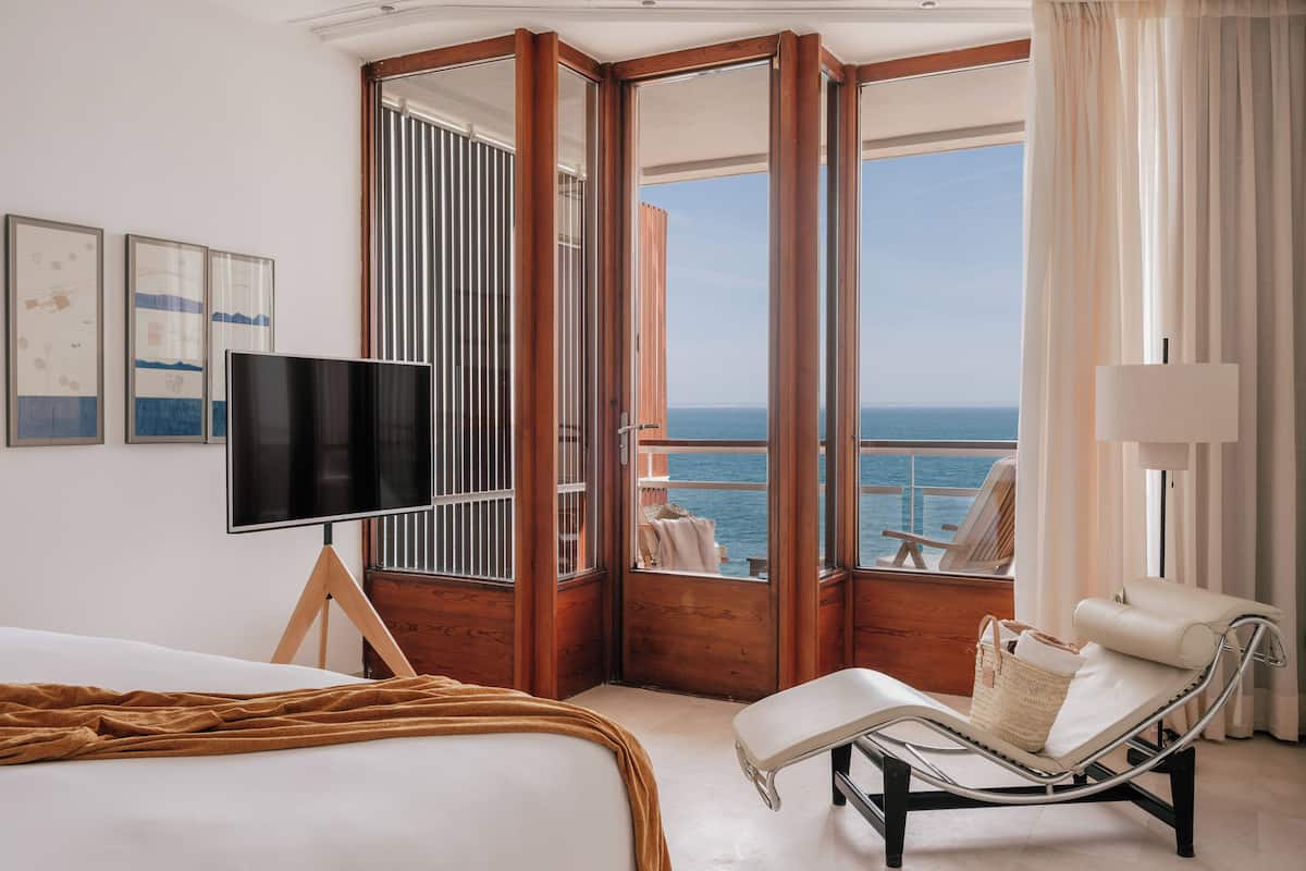 A hotel room with a bed, a TV, a lounge chair, and a balcony with a view of the sea.