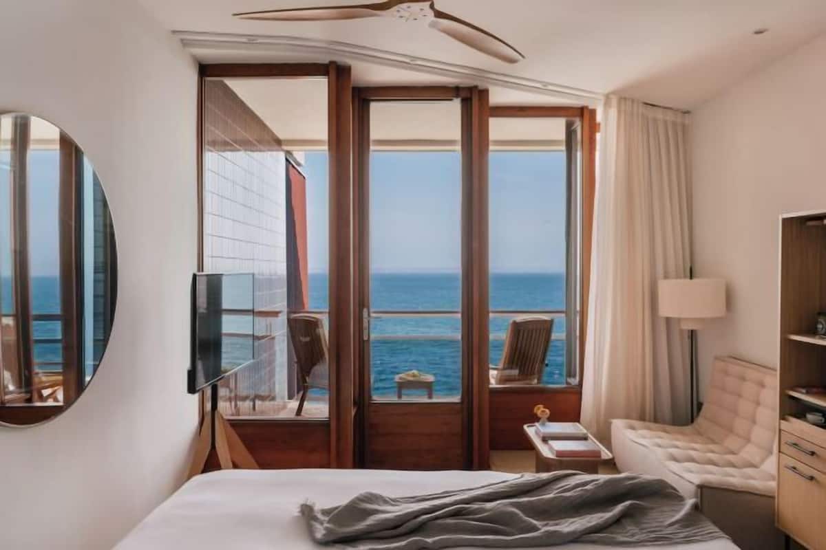 A hotel room with a bed, a TV, a desk, a chair, a lamp, and a view of the ocean.