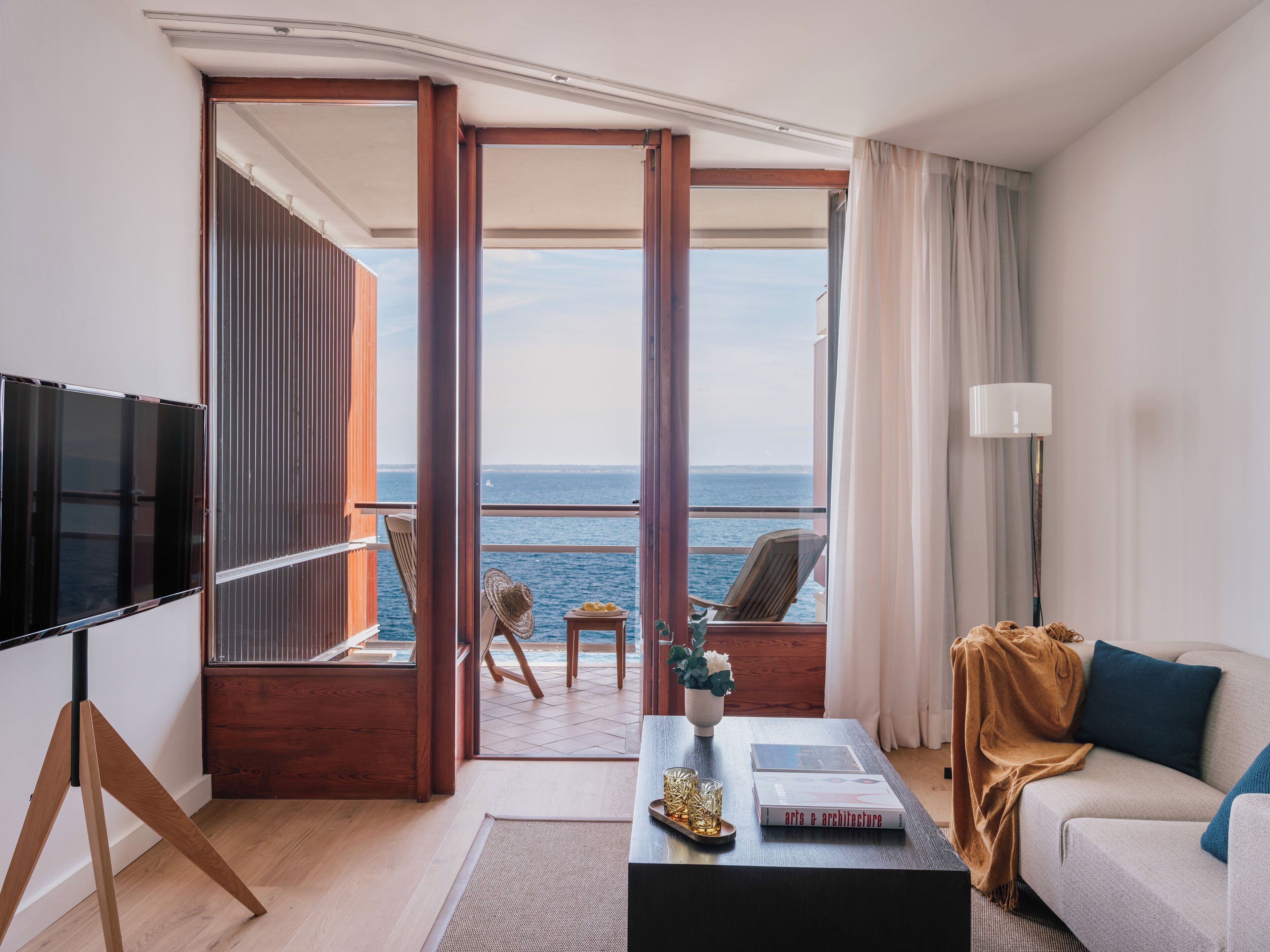 A modern hotel room with a balcony overlooking the sea, a sofa, a coffee table with books, and a TV.