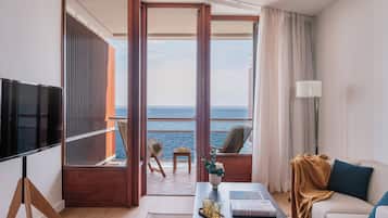 A modern hotel room with a balcony overlooking the sea, a sofa, a coffee table with books, and a TV.