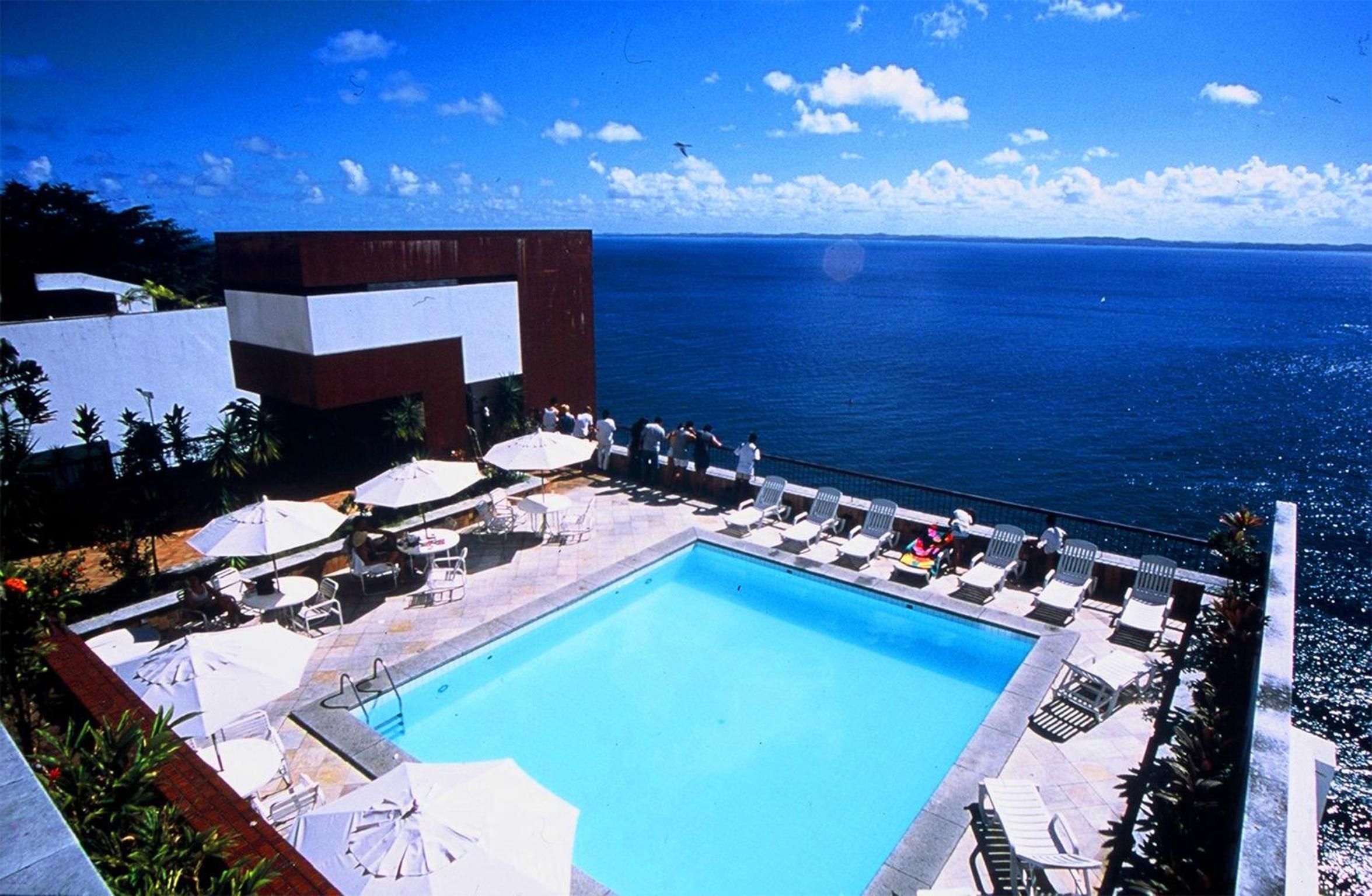 Zona de piscina con vista despejada al océano, rodeada de sillas de descanso y sombrillas.