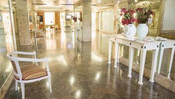 Un vestíbulo de hotel con piso de mármol, un mostrador de recepción, una silla blanca con cojín a rayas y un jarrón con flores.