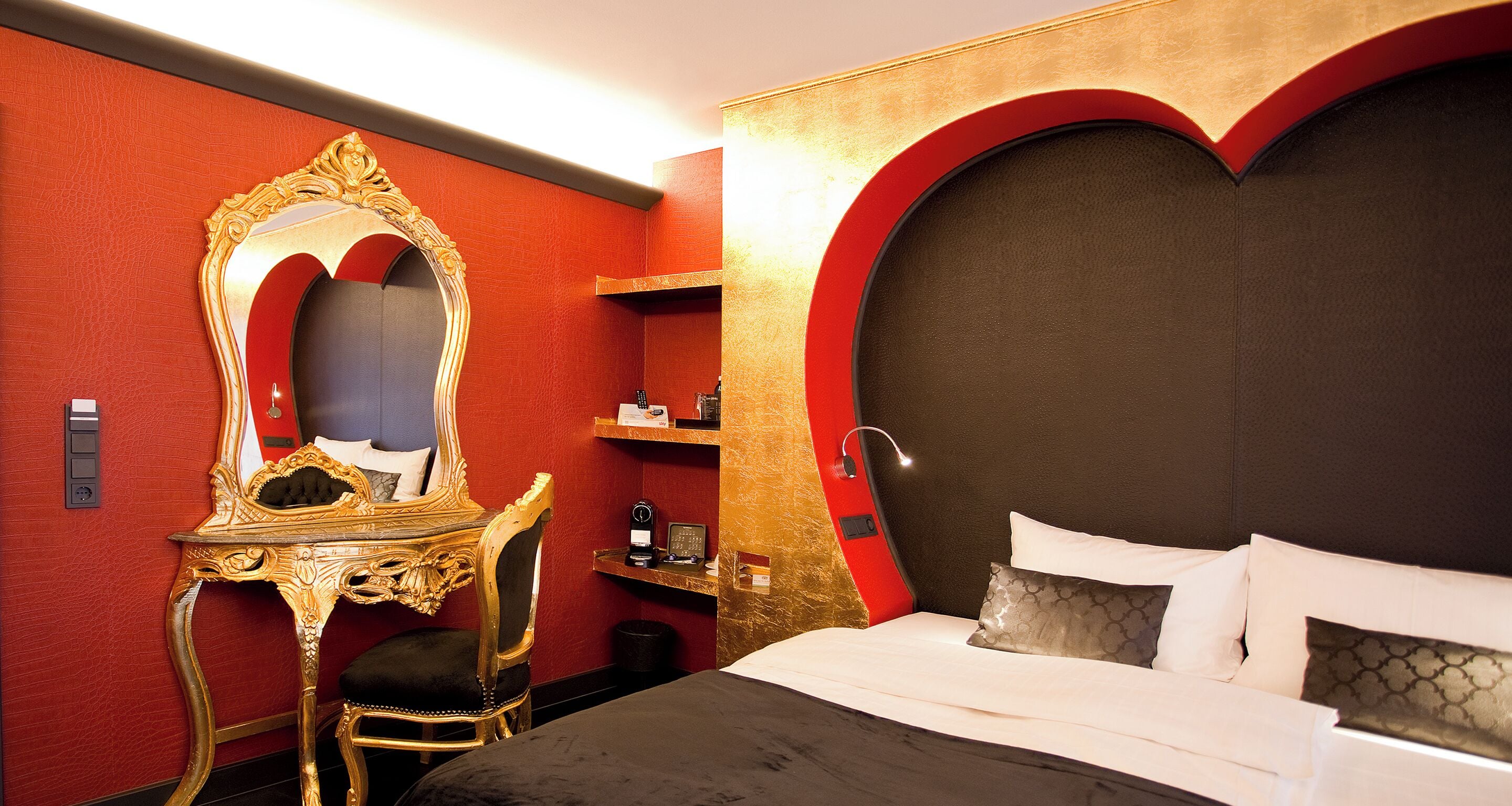 Ein Hotelzimmer mit einem großen Bett, einem spiegeln Spiegel mit goldener Rahmung, einem Schreibtisch und einer Wand in roter Farbe.