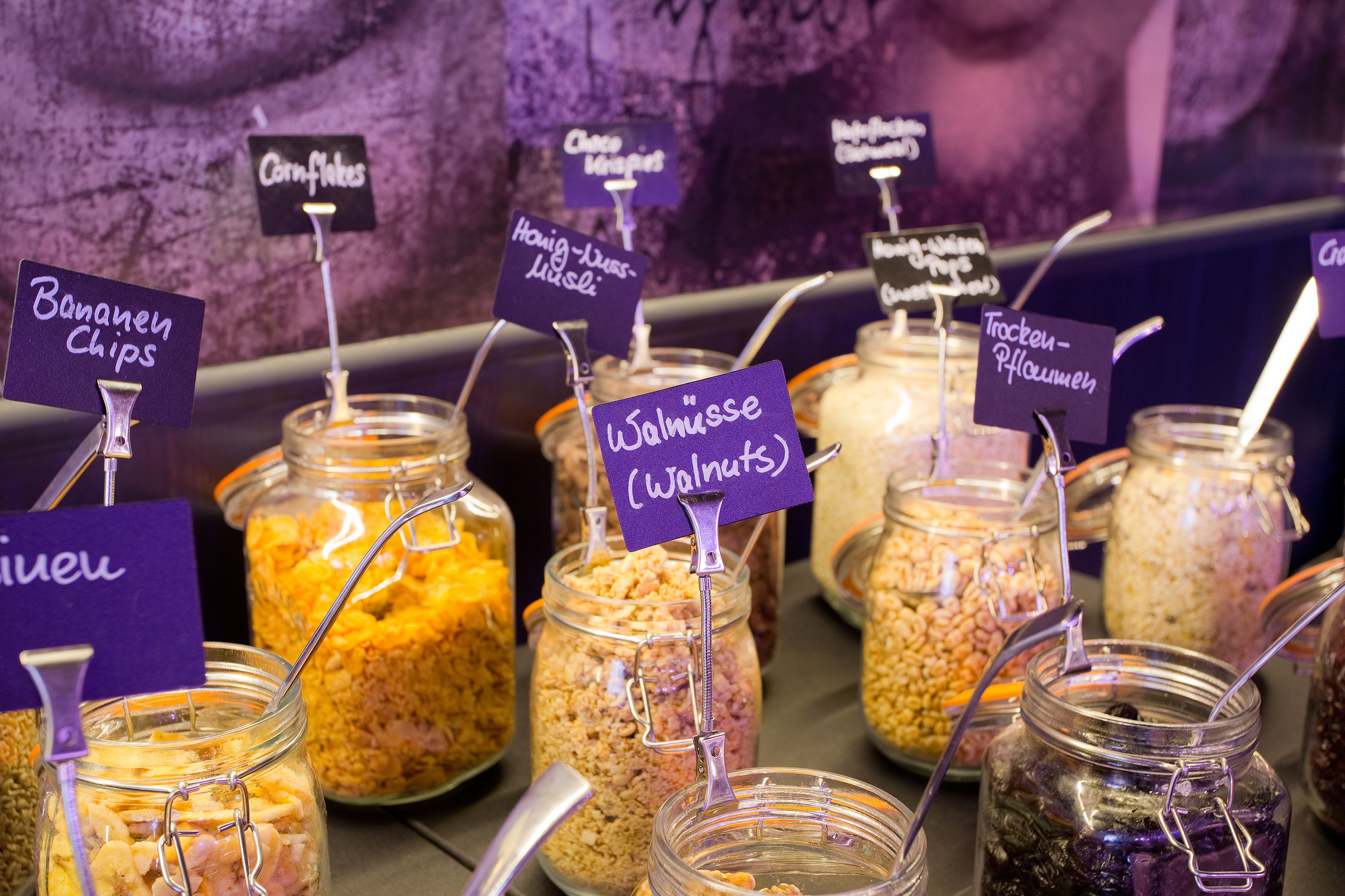 A variety of food items in jars with labels, including chips, cornflakes, cheese crisp, and walnuts.