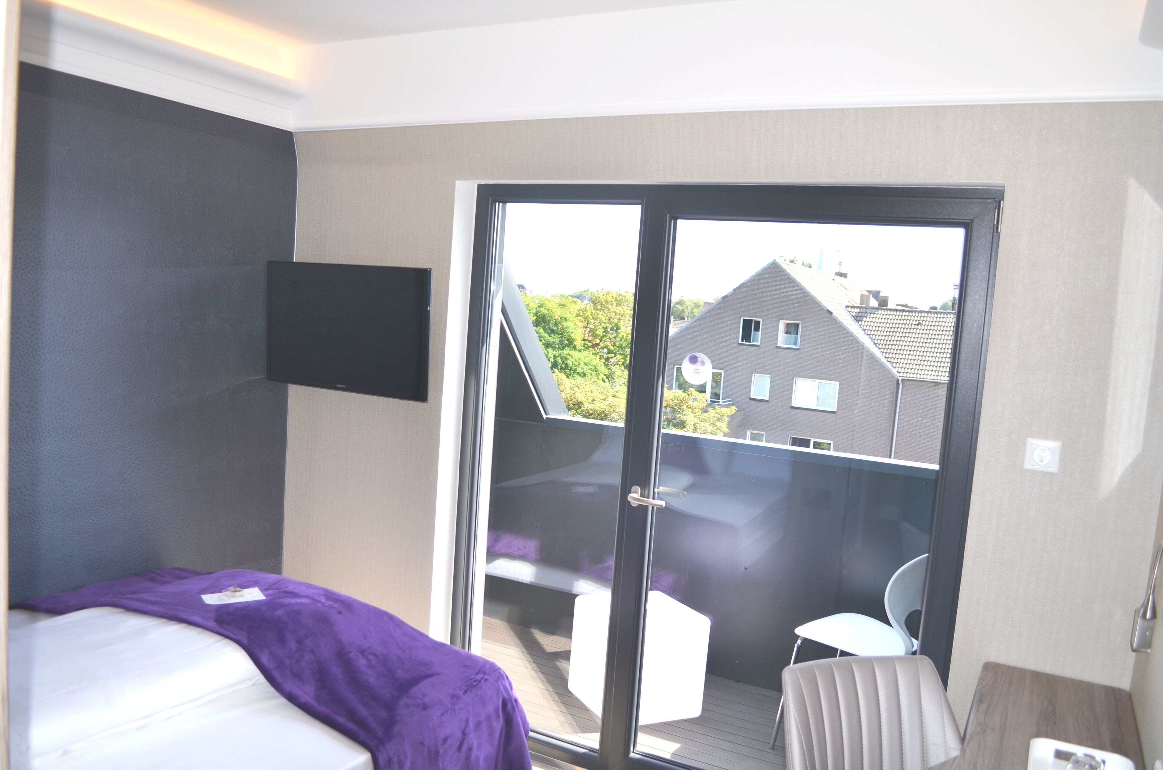 A hotel room with a bed, a TV mounted on the wall, a sliding glass door leading to an outdoor seating area, and a chair.