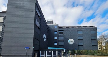 Un edificio moderno con una entrada destacada, grandes ventanales y un cartel que indica un área de negocios o servicios.