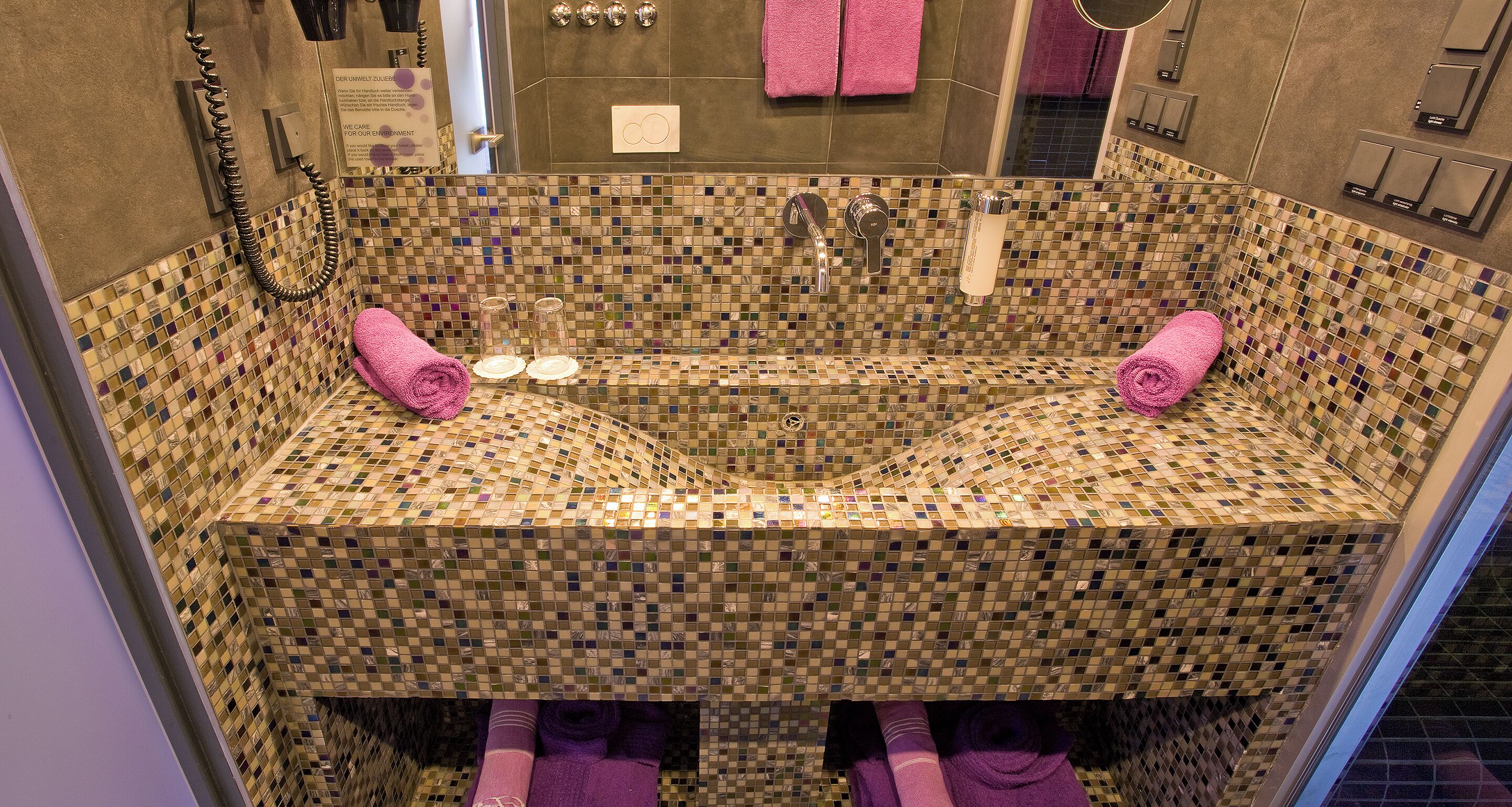 Ein Badezimmer mit Badewanne aus Mosaikfliesen, zwei rosa Handtüchern und einer Duschkabine mit Glastür.