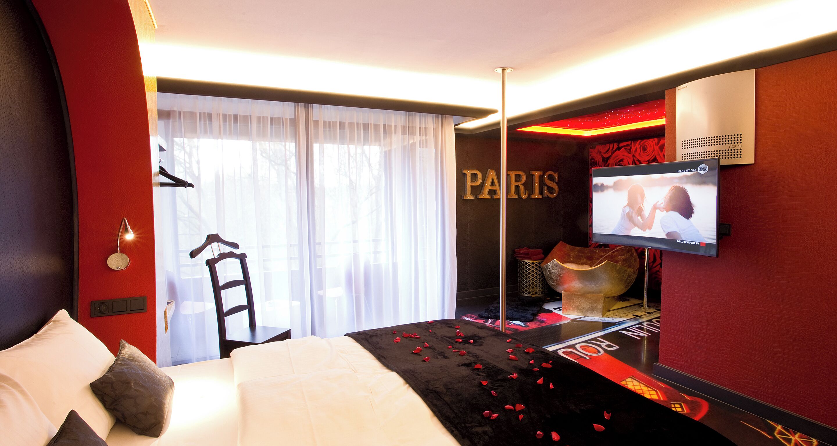 Une chambre d’hôtel avec un lit, une télévision, un mur rouge sur lequel est écrit le mot « PARIS », et une fenêtre avec des rideaux légers.