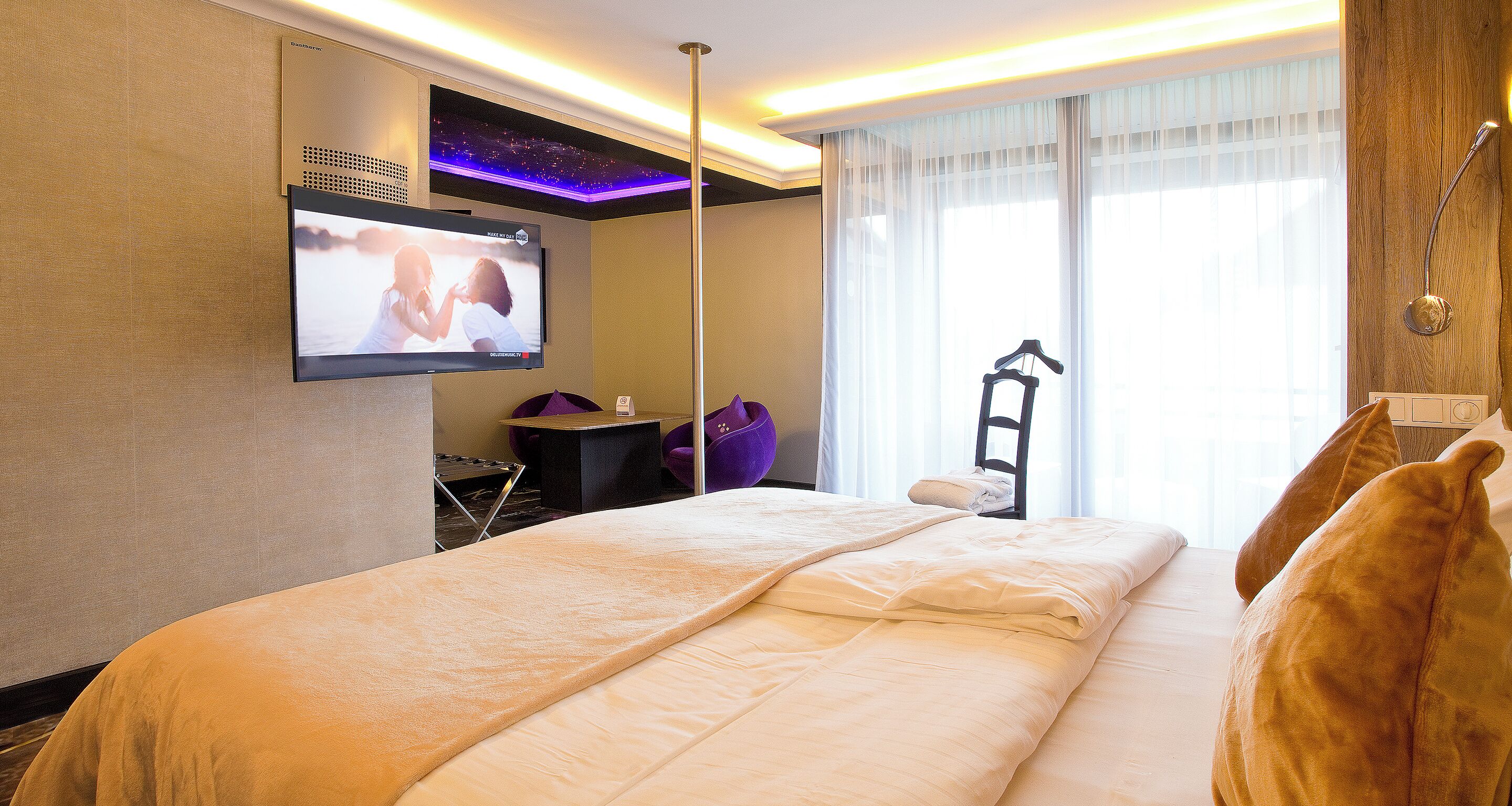 Ein Hotelzimmer mit einem großen Bett, einem flachbildfernseher an der Wand, einem Schreibtisch mit Stuhl und einem fenster mit Vorhängen.